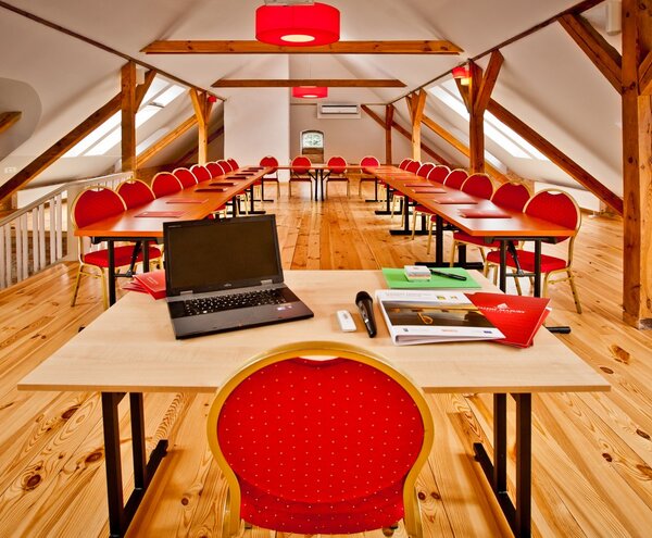 Sala konferencyjna na poddaszu z drewnianymi belkami, świetlikami, stołami w układzie U, czerwonymi krzesłami i biurkiem z laptopem i mikrofonem.