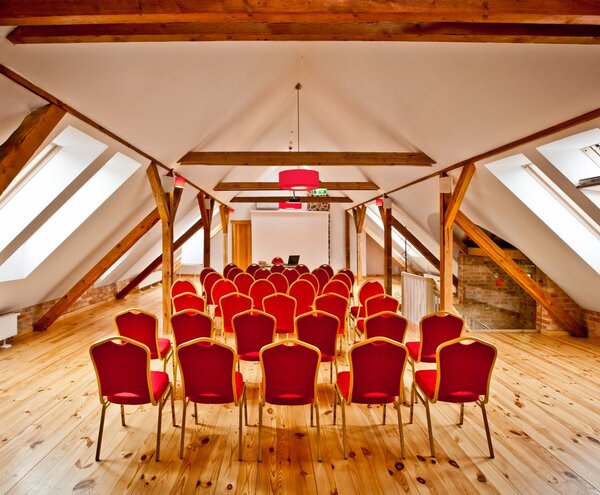 Sala konferencyjna na poddaszu z odsłoniętymi drewnianymi belkami, oknami dachowymi, polerowaną podłogą i czerwonymi krzesłami