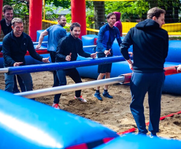 Goście grają w ludzkie piłkarzyki w dmuchanym, piaszczystym boisku — zewnętrzna atrakcja integracyjna z miękkimi drążkami i żywą atmosferą