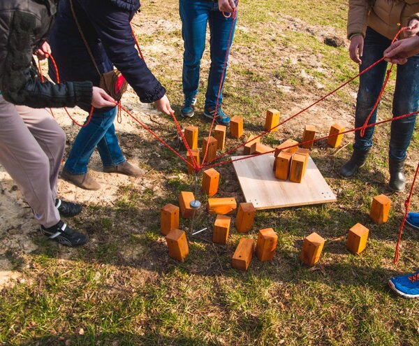 Goście w ubraniach codziennych stoją na trawie, trzymając czerwone liny, by wspólnie podnieść i ustawić drewniane klocki w grze integracyjnej.