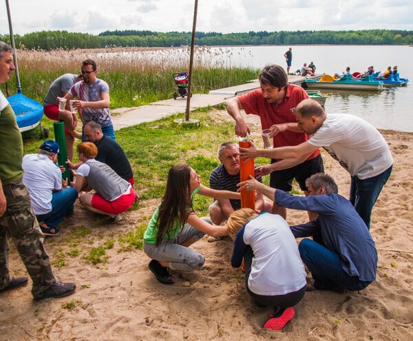 Warsztaty integracyjne nad jeziorem na plaży: goście współpracują przy pomarańczowej rurze, w tle pomost i rowerki wodne.