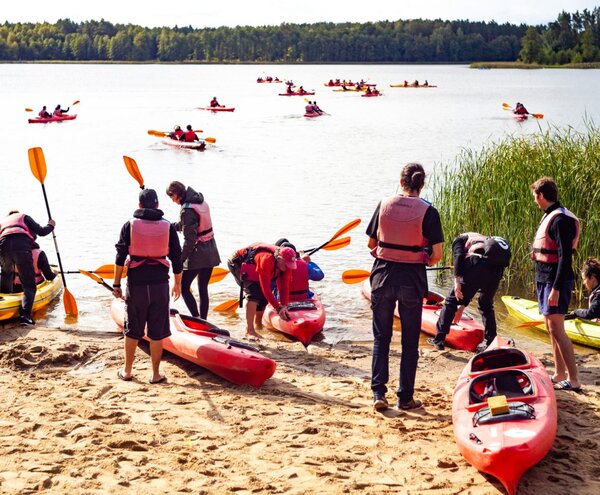 Goście w kamizelkach wypływają czerwonymi i żółtymi kajakami z piaszczystej plaży, płynąc po spokojnym jeziorze przy zalesionym brzegu.