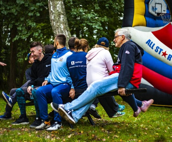 Grupa gości balansuje na długiej huśtawce-ważnicy na trawniku podczas gry integracyjnej obok dmuchanej ścianki do koszykówki.