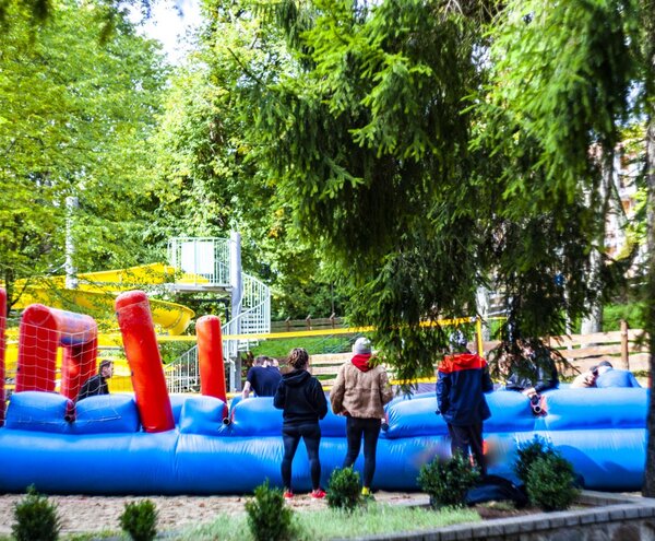 Niebieska nadmuchiwana przeszkoda z czerwonymi słupami, goście obok, żółta spiralna zjeżdżalnia i leśny park.