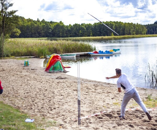 Piaszczysta plaża nad jeziorem z łódkami pedałowymi i kolorowymi kajakami; gość rzuca kijem do gry, instruktor obserwuje.