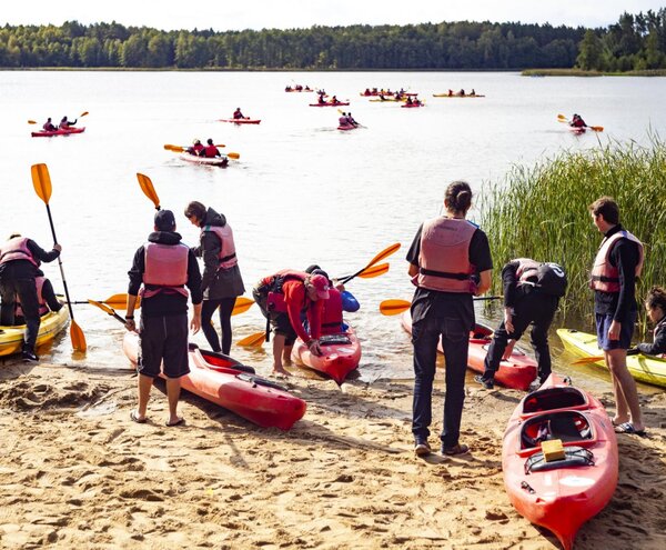 Grupowy spływ kajakowy z piaszczystej plaży, goście w kamizelkach wypływają czerwonymi i żółtymi kajakami po spokojnym jeziorze
