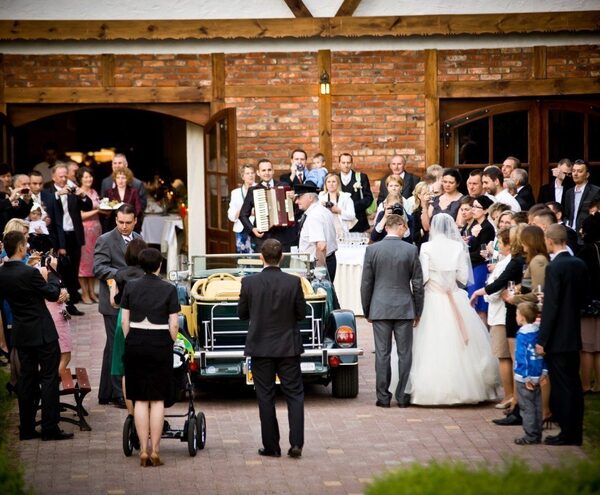 Wedding reception in rustic brick courtyard: bride and groom by vintage car, guests mingling, live accordion and drinks.