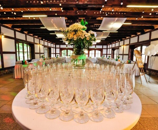 Rustic banquet hall with wooden beams, draped ceiling lights, long set tables, rows of champagne glasses and a central floral centerpiece.