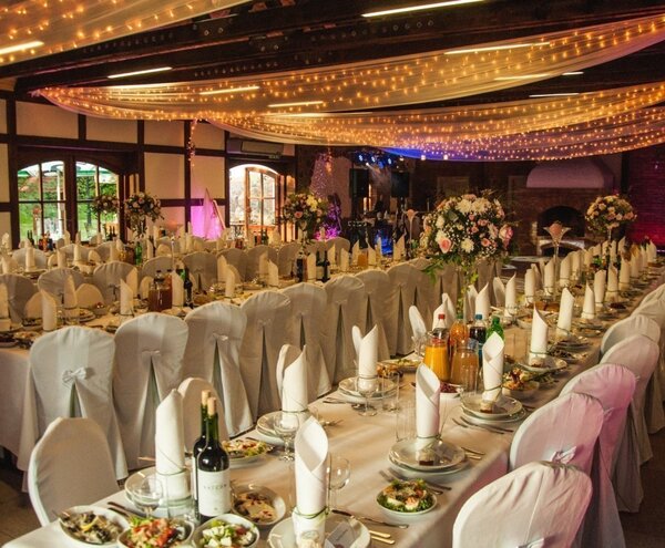 Elegant banquet hall set for a wedding: long white-covered tables with folded napkins, floral centerpieces, wine and a fairy-light draped ceiling.