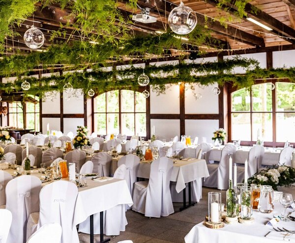 Rustic reception hall with white-covered chairs, long set tables, suspended greenery, glass orbs and floral centerpieces.