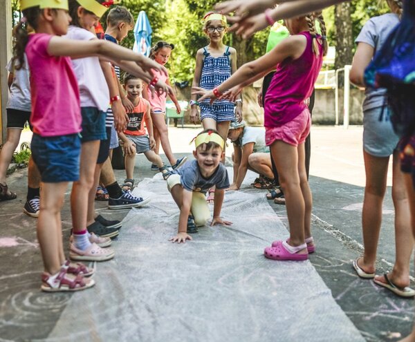 Kids at an outdoor hotel kids' club crawl through a human tunnel on a chalk-covered courtyard mat during summer activities