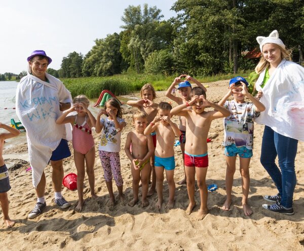 Children and two entertainers making heart hands on a sandy lakeside hotel beach during supervised summer fun