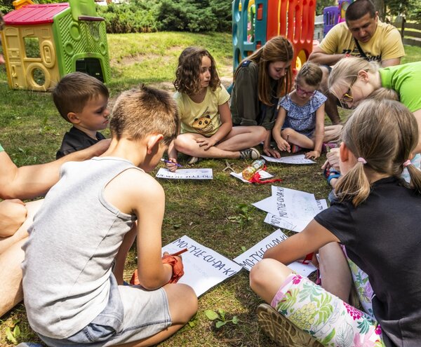 Children and staff at a hotel's supervised outdoor kids' club, seated on grass doing arts-and-crafts near colorful play structures.
