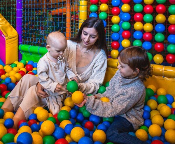 Family-friendly indoor kids' club ball pit with colorful balls and netted soft-play walls; mother supervising two children playing.