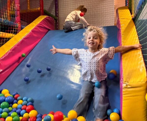 Hotel indoor playroom: soft slide into vibrant ball pit, child sliding with arms out, another climbing back up.