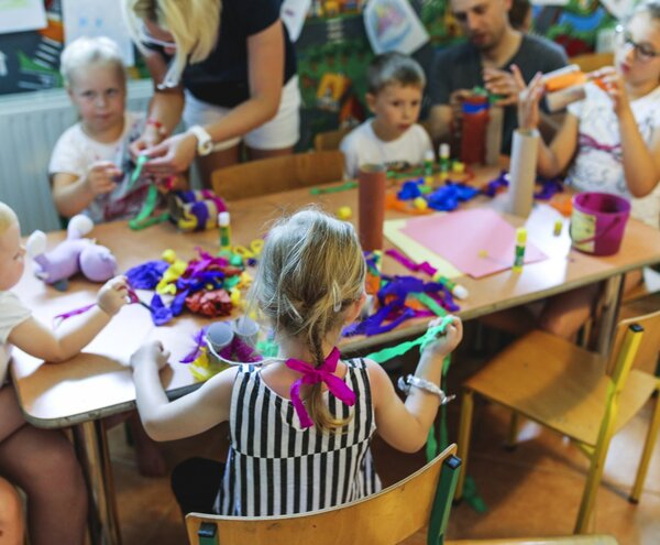 Children and a caregiver at a hotel kids' club table making colorful crafts with ribbons, cardboard tubes and glue sticks.