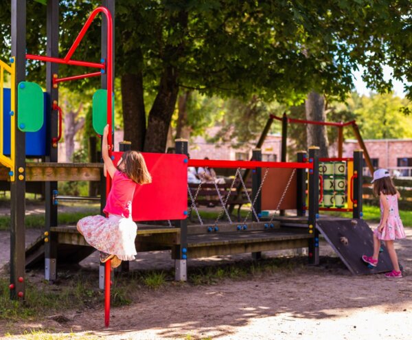 Plac zabaw w cieniu z kolorową konstrukcją do wspinaczki, rurą i pochyloną ścianką; dwie dziewczynki bawią się przy drzewach obok budynku hotelowego.