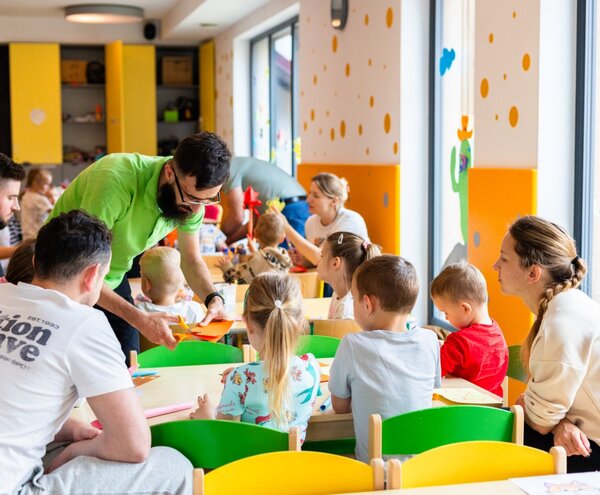 Jasny klub dziecięcy w hotelu z dużymi oknami, kolorowymi krzesłami, stołami do zajęć i animatorem pomagającym dzieciom.