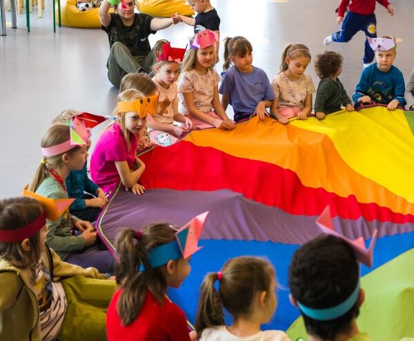 Hotel children's club in playroom: kids and an animator sit in a circle holding a large rainbow parachute, wearing paper crowns.