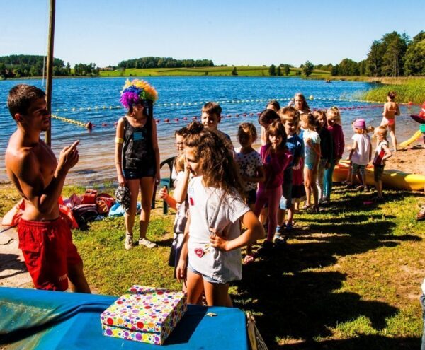 Lakeside family activity on a supervised beach: children lined up for games led by staff near a roped swim area and kayaks.