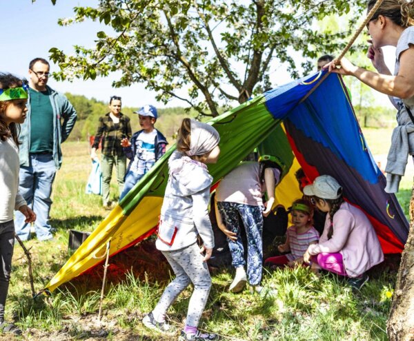 Children playing under a colorful parachute-style tent tied to a tree in a sunny meadow, adults supervising nearby