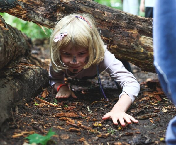Blond child with face paint crawling barefoot under a fallen log on a forest floor, playful outdoor adventure.