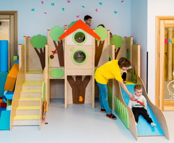 Supervised indoor kids' playhouse with wooden tree-themed climb, slide and staff assisting toddlers