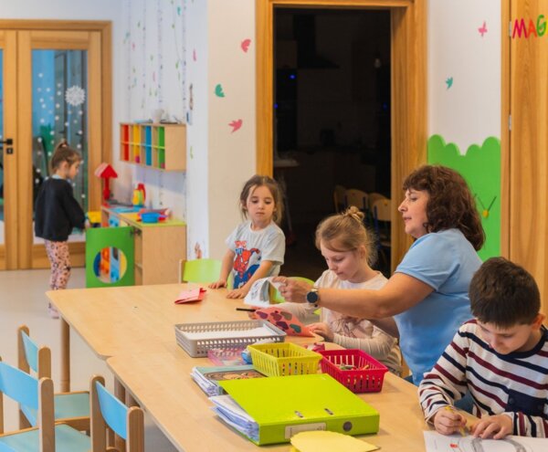 Bright children's activity room with wooden tables and colorful shelves; caregiver guiding children in arts and crafts and drawing.
