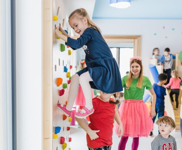 Children's playroom with supervised kids' club: girl climbing colorful indoor climbing wall, staff leading games and soft-play area.