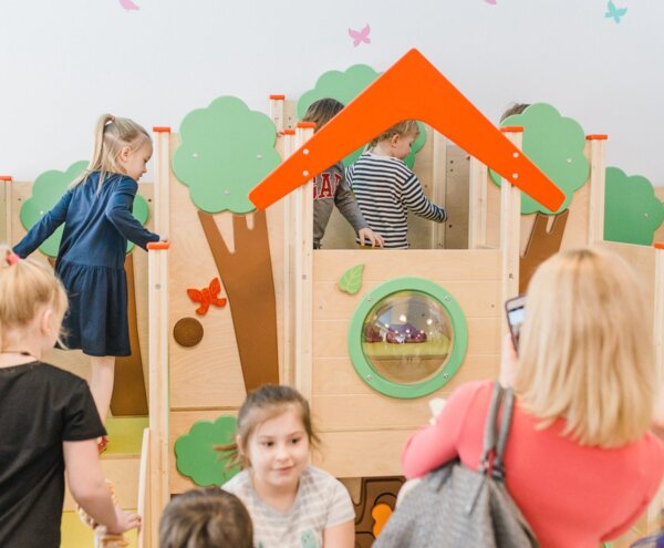 Children playing in a wooden indoor playhouse with green tree panels, orange roof and bubble window while adults watch and take photos.