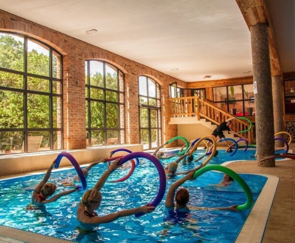 Kryty basen hotelowy przy łukowych ceglastych oknach; grupa aqua fitness z kolorowymi makaronami i instruktorem.