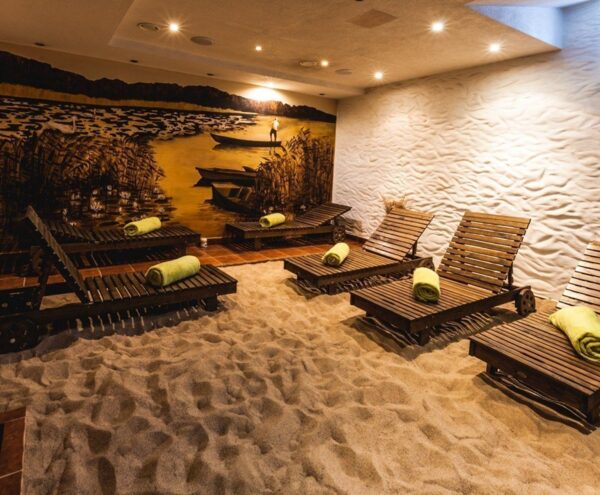 Indoor spa relaxation room with sand floor, wooden lounge chairs each with rolled green towel, warm mural of boats and reeds.