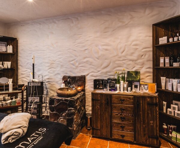 Cozy spa treatment room with warm lighting, rustic wooden cabinets, stone basin, shelves of skincare products and a treatment bed with folded towels.