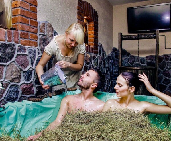 Couple relaxing in a hay-filled spa tub as attendant pours water from a metal bucket in a rustic stone and brick treatment room.
