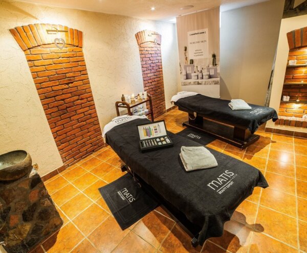 Couples spa treatment room with two massage tables, Matis towels, skincare display, warm tile floor and exposed brick arches.