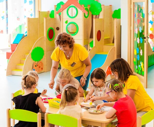 Hotel kids' club craft session with two staff supervising children at a table, colorful playhouse and indoor slides behind