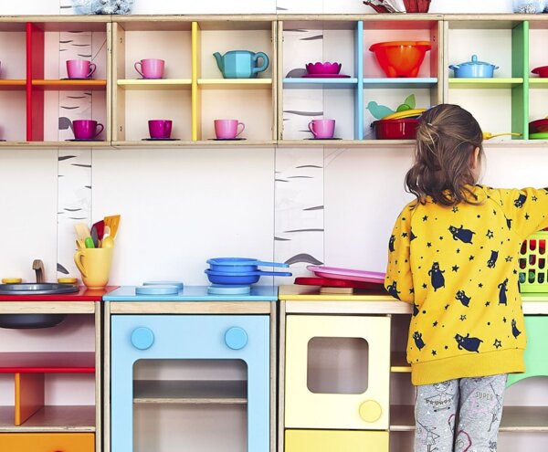 Family-friendly kids' play corner with wooden colorful toy kitchen, cups, pans, small chairs and wall birch decals; child playing.