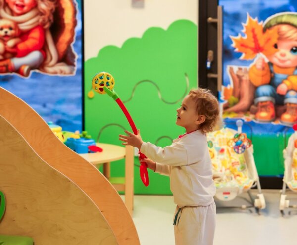 Toddler playing with a colorful rattle in a bright hotel kids' playroom with murals, activity table and baby bouncers.