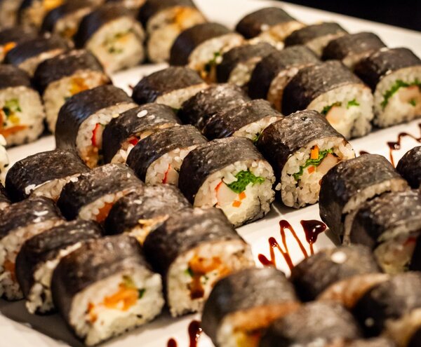 Assorted nori-wrapped sushi rolls with rice, vegetable and crab fillings, arranged on a hotel buffet platter with drizzled sauce.