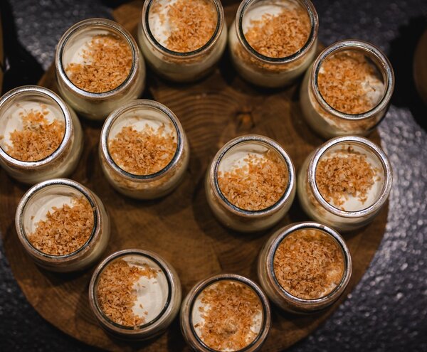 Individual creamy desserts in glass jars topped with toasted coconut, arranged on a wooden board at a buffet/dessert station
