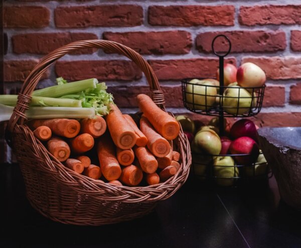 Breakfast produce display: wicker basket of carrots and celery beside a two-tier wire stand of apples against brick wall