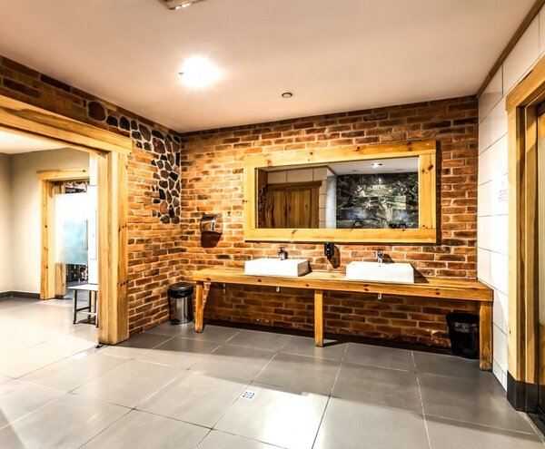 Łazienka SPA w stylu rustykalnym: cegła, duże drewniane lustro, dwie umywalki na drewnianym blacie i wejście do sauny.
