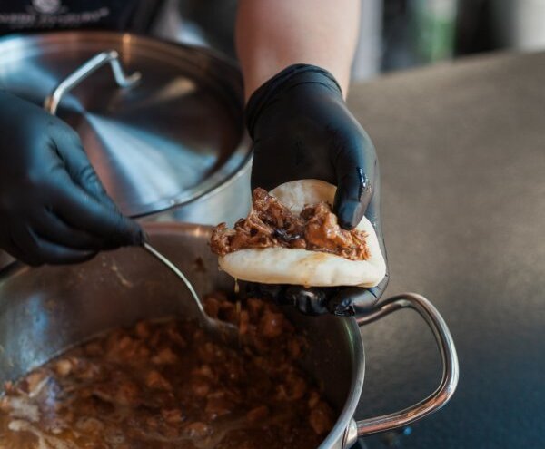 Chef scooping slow-braised pulled meat into a steamed bun over a stainless pot, hands in black gloves in open kitchen.
