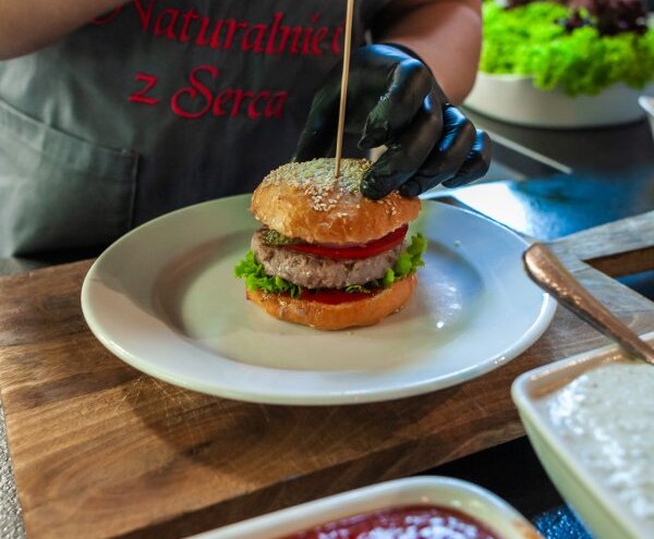 Chef assembling gourmet burger on white plate, sesame bun with lettuce, tomato and patty, sauces and salad bowl nearby