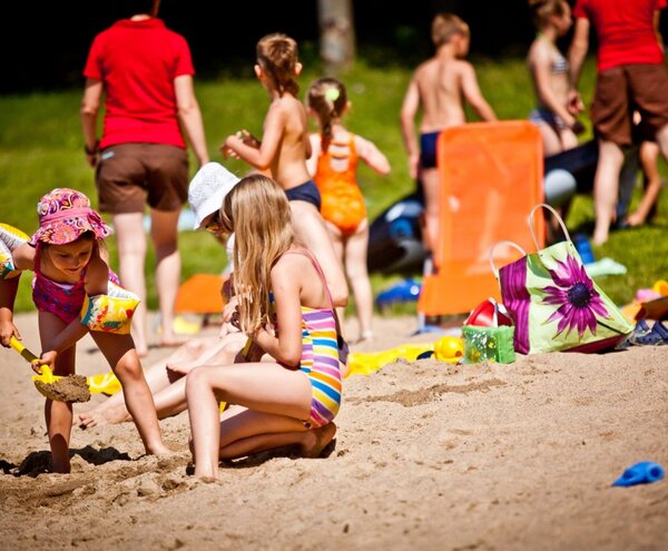 Family-friendly sandy beach with children digging in sand, colorful lounge chairs, inflatable armbands and a floral beach bag nearby.