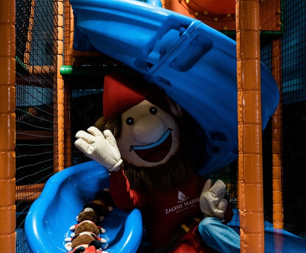 Indoor children's play area with blue spiral slide, padded climbing frame and a waving hotel mascot plush toy.