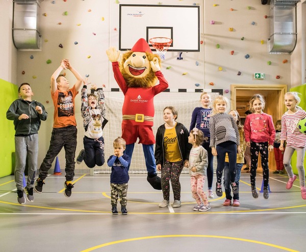 Children playing with a hotel mascot in a bright indoor activity hall with climbing wall, basketball hoop and padded walls.