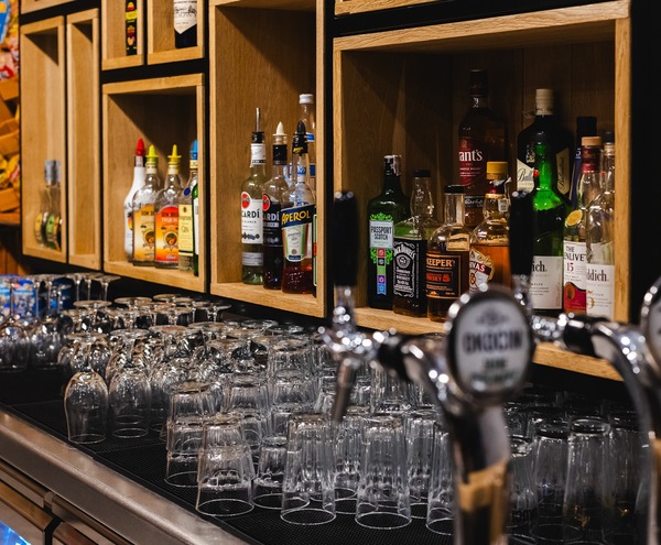 Bar hotelowy z drewnianymi półkami pełnymi alkoholi, krany piwne na pierwszym planie i rzędem czystych kieliszków.