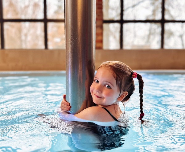 Smiling girl in a hotel indoor pool hugging a metal column, braided hair with flower clip, large grid windows and calm water