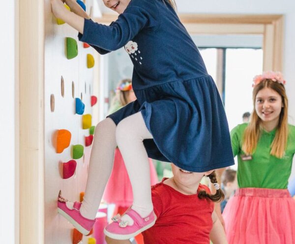 Hotel kids' club: child climbing a colorful indoor wall while other children play and a staff member supervises.